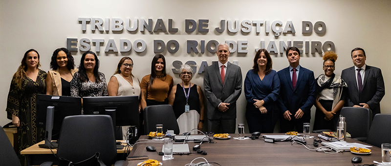 Foto de grupo, homens e mulheres, que participou de reunião de reunião sobre salas de apoio à amamentação. Todos estão atrás de uma mesa e à frente de letreiro do Tribunal de Justiça do Estado do Rio de Janeiro