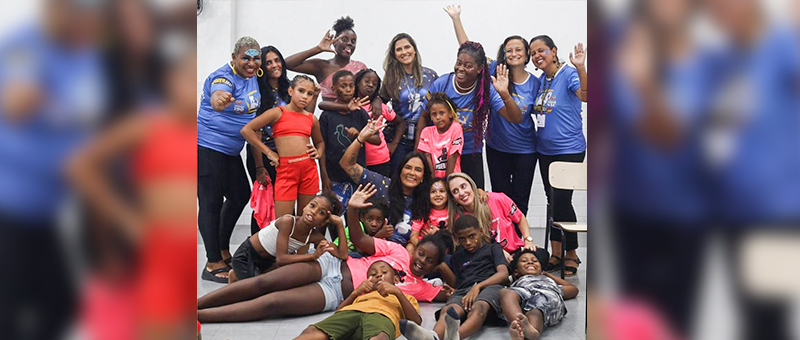 Sete recreadoras de camisa azul e calça preta posam para foto com grupo de crianças sentadas e deitadas no chão, vestindo roupas coloridas, em uma sala de paredes claras 