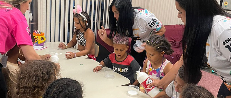 Oito crianças brincam ao redor de uma mesa com apoio de três educadoras em sala adaptada para a recreação infantil de forma temporária