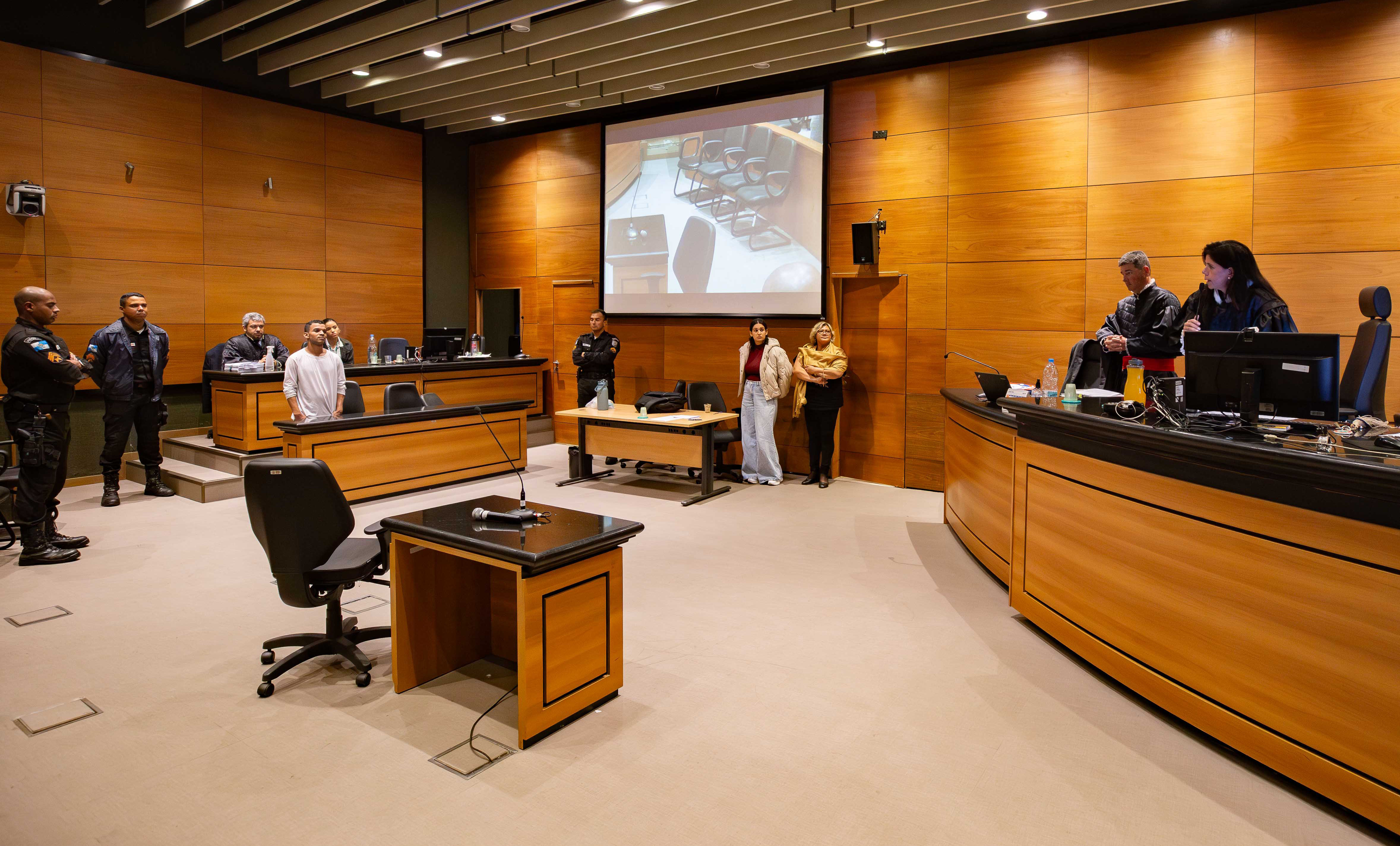 Imagem mostra cena de um julgamento em Tribunal do Júri com o réu, um homem, de pé, ouvindo a sentença que terá de cumprir sendo proferida pela juíza