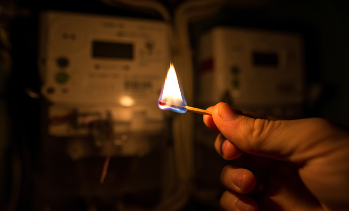 #Paratodosverem: A imagem mostra uma mão segurando um fósforo aceso em primeiro plano, com foco na chama. O fundo está desfocado, mas revela dois medidores de energia elétrica, com luz ambiente baixa ou ausência de iluminação elétrica.